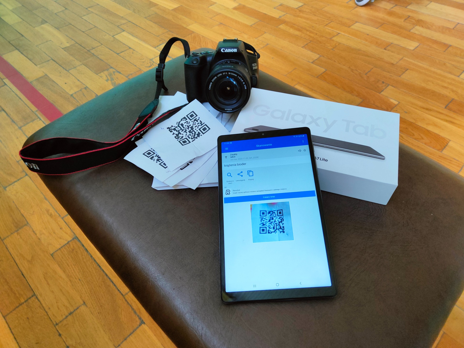 Na zdjęciu znajduje się aparat fotograficzny, kartki z kodami QR oraz tablet Samsung.