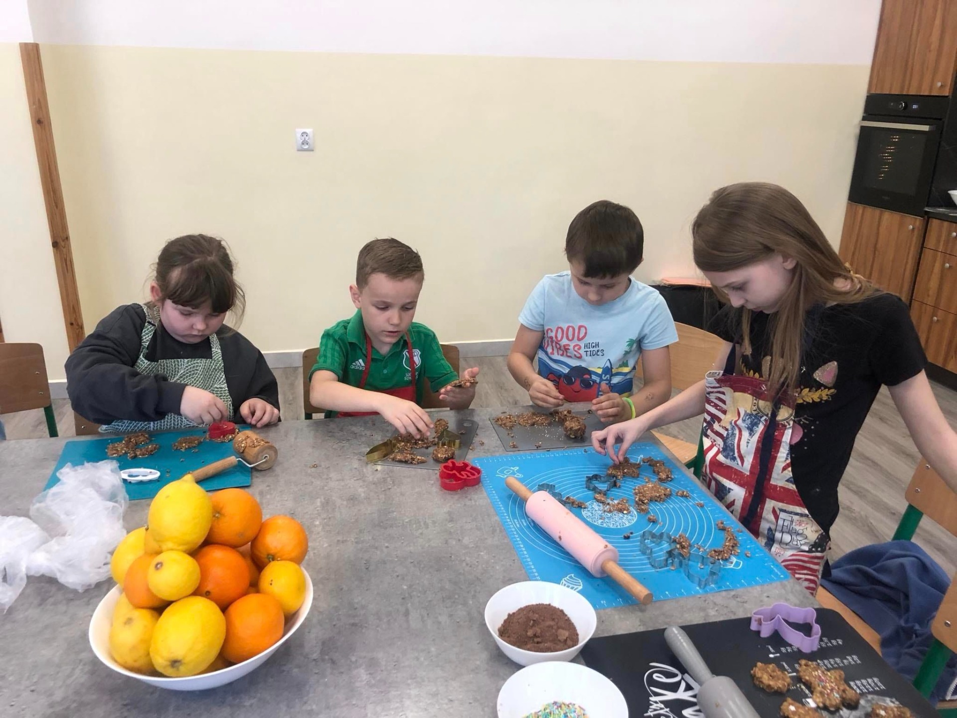 Na zdjęciu widzimy grupę dzieci siedzących przy długim stole w jasnym pomieszczeniu. Dzieci są zaangażowane w tworzenie rękodzieła lub dekoracji.