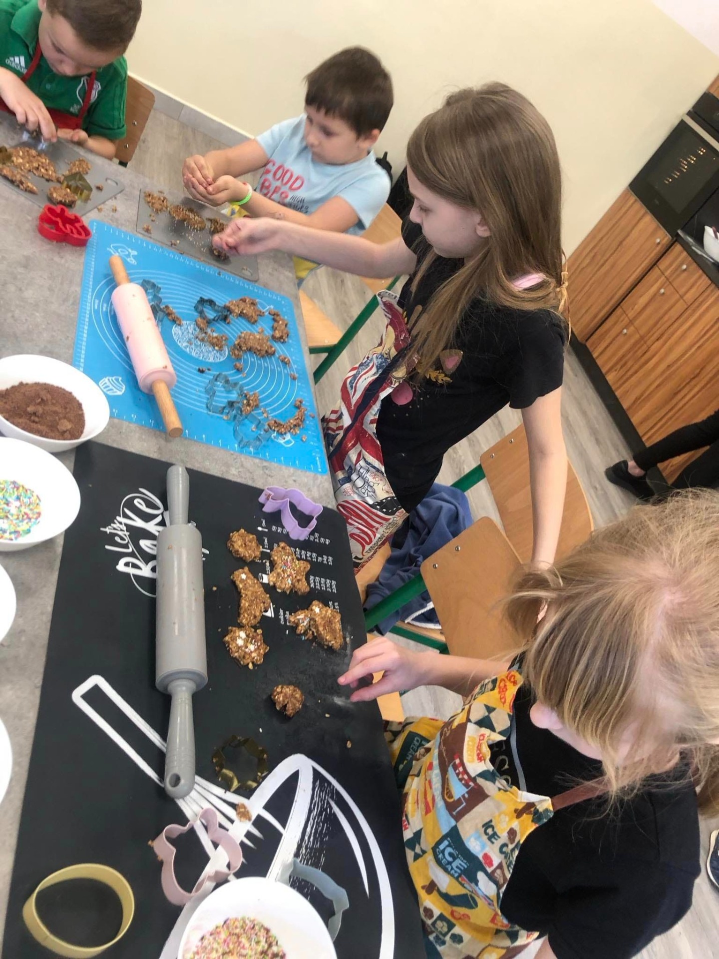 Na zdjęciu widzimy grupę dzieci zaangażowanych w wspólne pieczenie ciastek. Siedzą przy stole, a przed nimi rozłożone są różne narzędzia i składniki do wypieku.