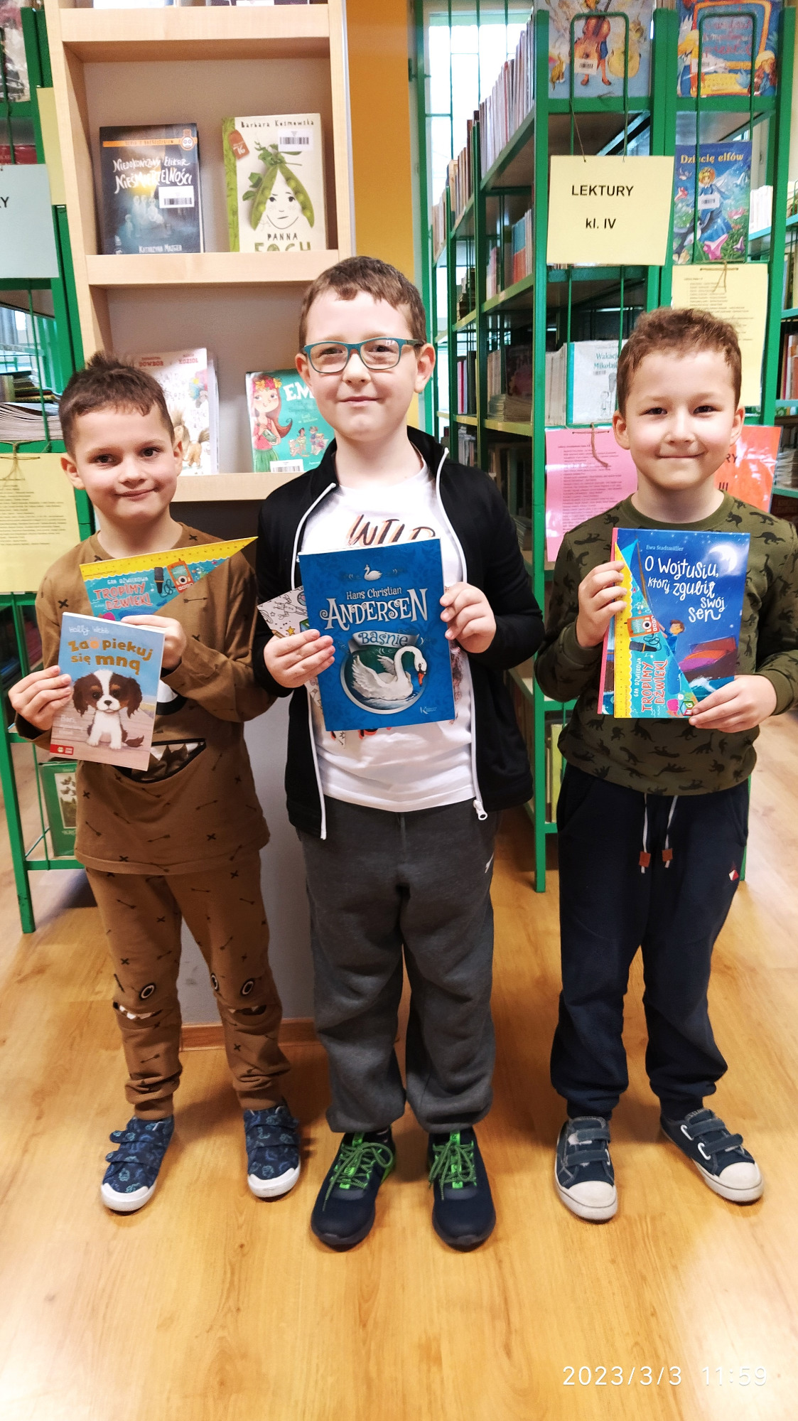 Troje uczniów w bibliotece szkolnej stojących przy półkach z książkami. Każdy z chłopców trzyma w dłoniach nagrodę książkową, którą prezentują.