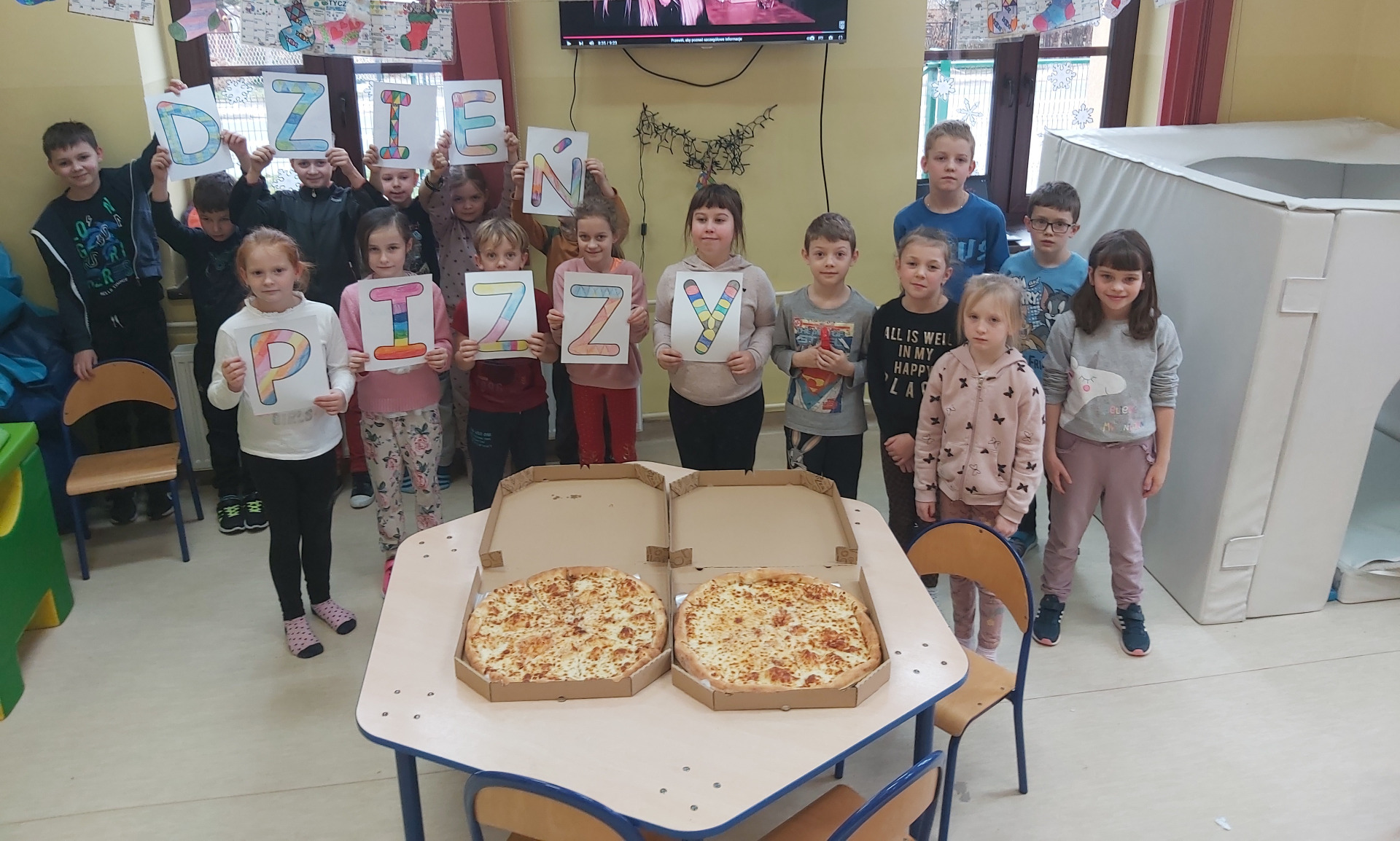 Dzieci ze świetlicy stoją wokół stołu, na którym leżą dwie pizze, niektóre z nich trzymają napis Dzień Pizzy