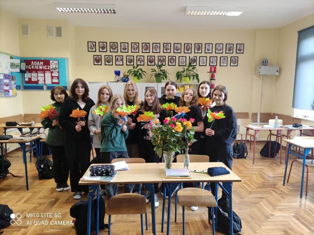 Na czwartym zdjęciu jest dziesięć uczennic klasy ósmej a trzymających w ręce papierowe kwiaty. Przed nimi na ławce stoi wazon z różami. W tle kolorowe gazetki, szafki na których znajdują się doniczki z kwiatami. Nad szafkami wiszą portrety polskich pisarzy.