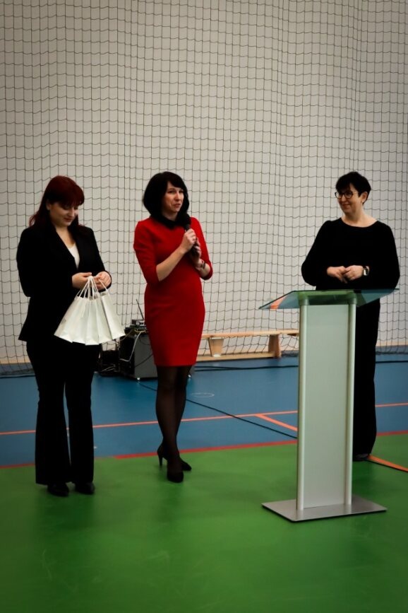 Zdjęcie z apelu na sali gimnastycznej:  pani dyrektor z dwiema przedstawicielki firmy CCS z nagrodami dla uczniów.