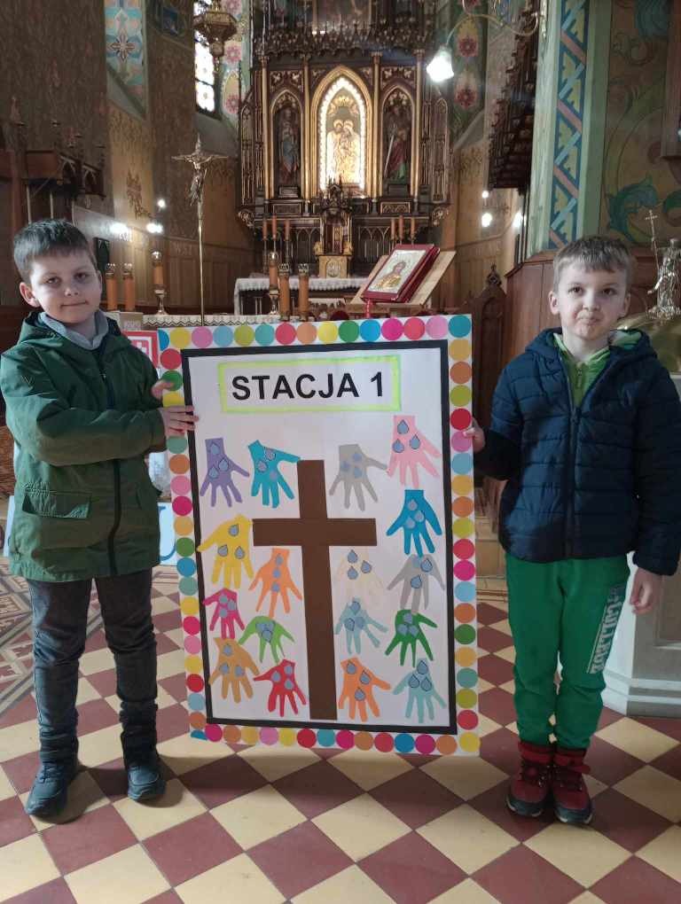 Na zdjęciu dwóch uczniów trzyma plakat przedstawiający pierwszą stację drogi krzyżowej.
