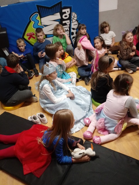 Dzieci siedzą na podłodze, a jedna dziewczynka leży na materacu i trzyma maskotkę.