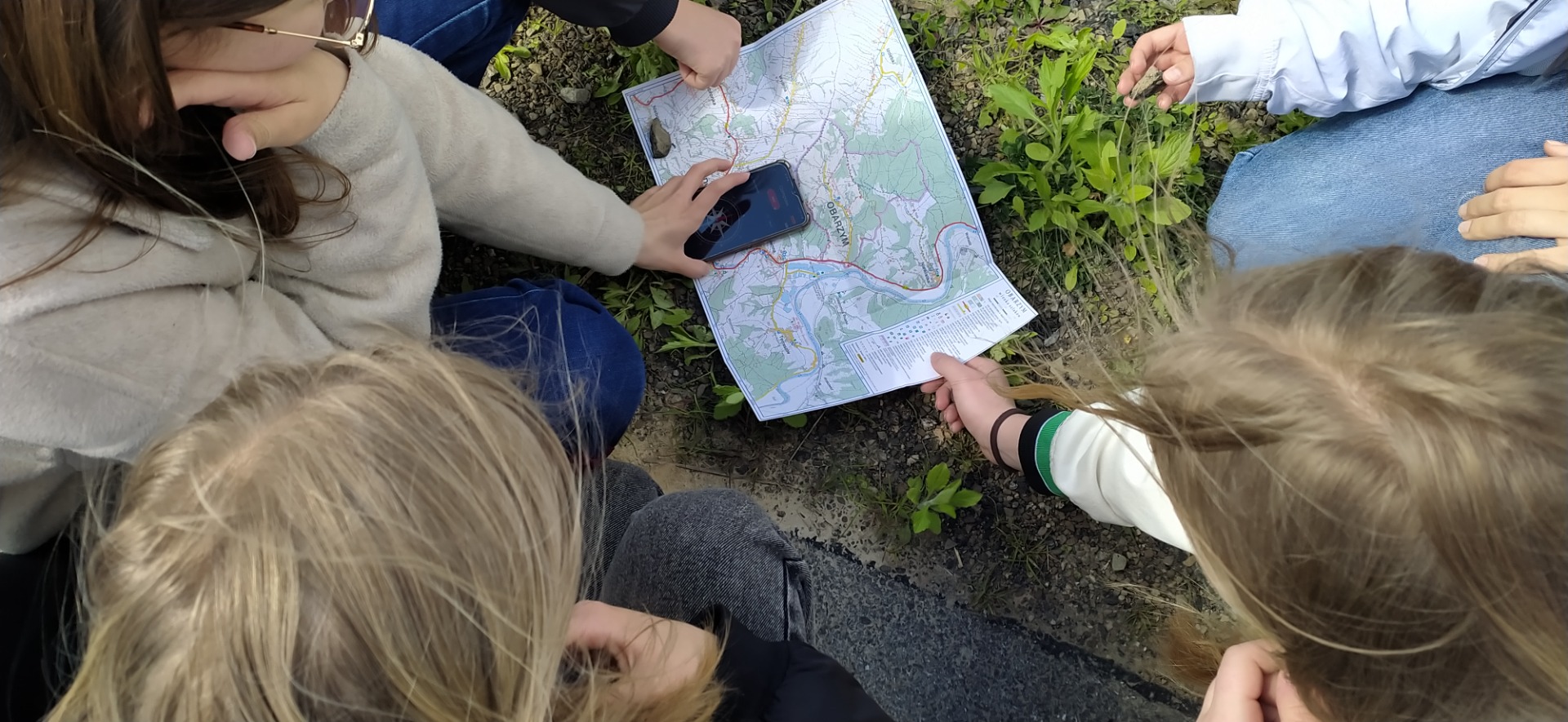 Na fotografii widać leżącą rozłożoną na trawie mapę gminy Dydnia, uczennica trzyma telefon z aplikacją kompasu.