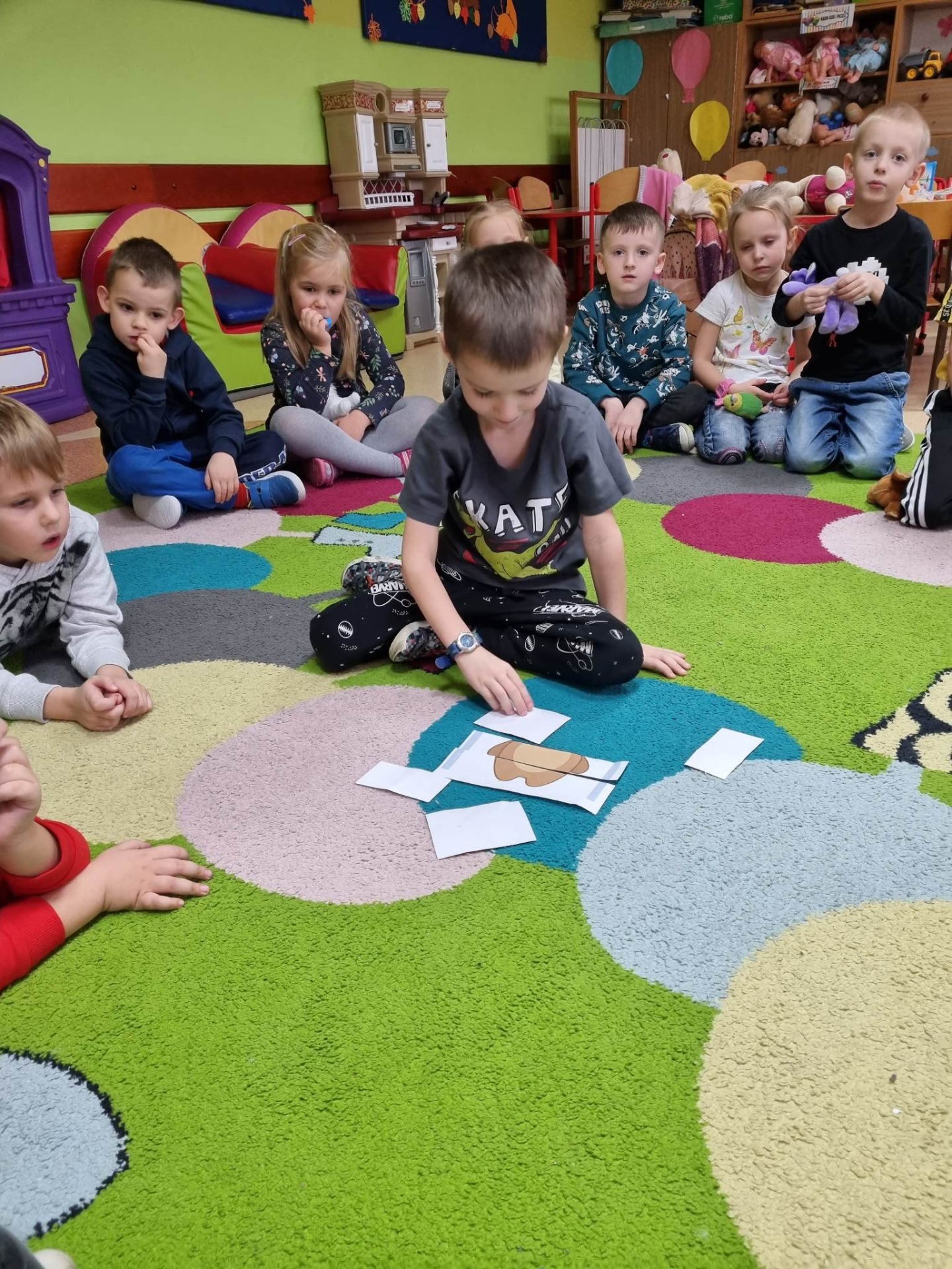 Grupa przedszkolaków siedzi w kole na kolorowym dywanie i rozwiązuje misiowe gry