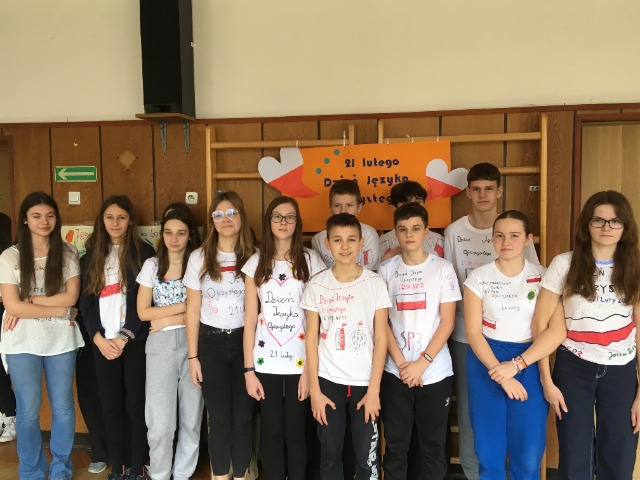 12 uczniów stoi ubranych w białe koszulki, na których narysowali symbole związane z Międzynarodowym Dniem Języka Ojczystego. Nad nimi wisi pomarańczowy plakat z napisem Międzynarodowy Dzień Języka Ojczystego.