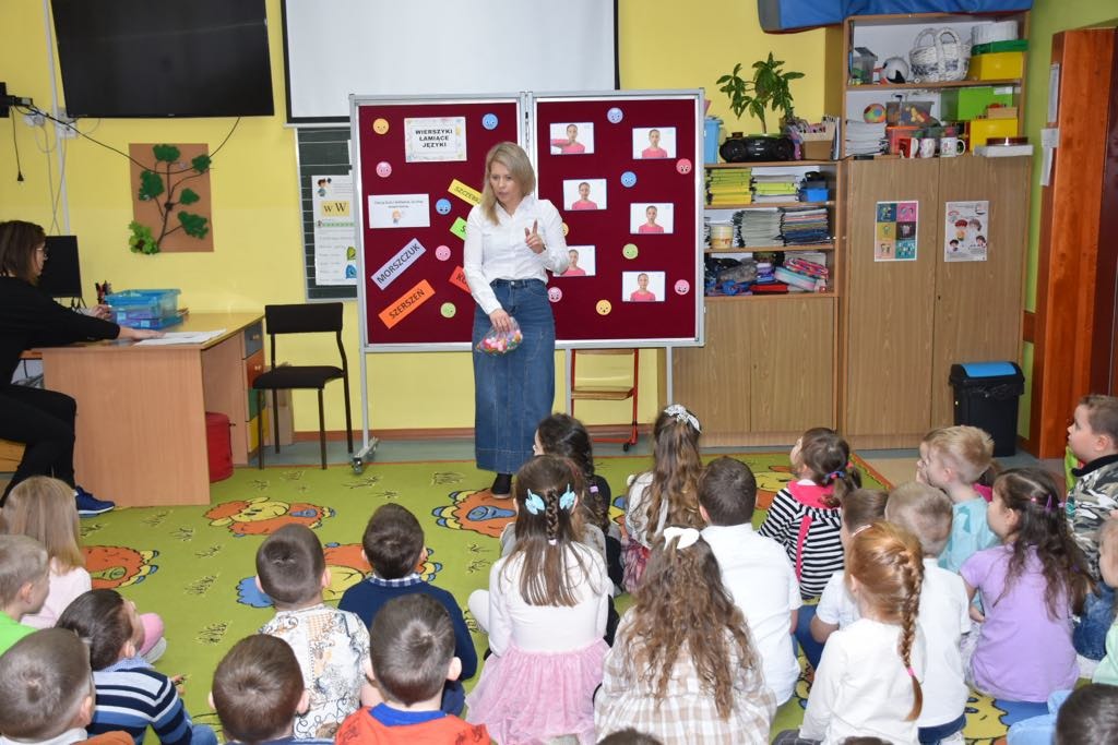 Nauczycielka stoi na tle gazetki tematycznej z napisem "Wierszyki łamiące języki" i opowiada dzieciom o celach konkursu.