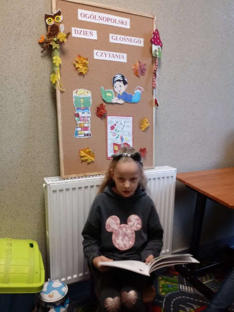 Dziewczynka siedzi i na kolanach trzyma otwartą książkę. Za nią tablica korkowa z napisem Ogólnopolski Dzień Głośnego Czytania.