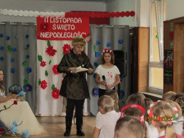 Dwoje uczniów stoi na scenie. Jeden uczeń przebrany za Tadeusza Kościuszkę, ma na sobie palto i czapkę z piórem. Uczennica ma wianek na głowie.
Nad nimi wisi czerwony plakat z białym napisem 11 listopada Święto Niepodległości.

