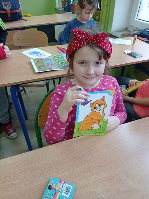 W ławce szkolnej siedzi dziewczynka, trzyma przed sobą rysunek z kotkiem.