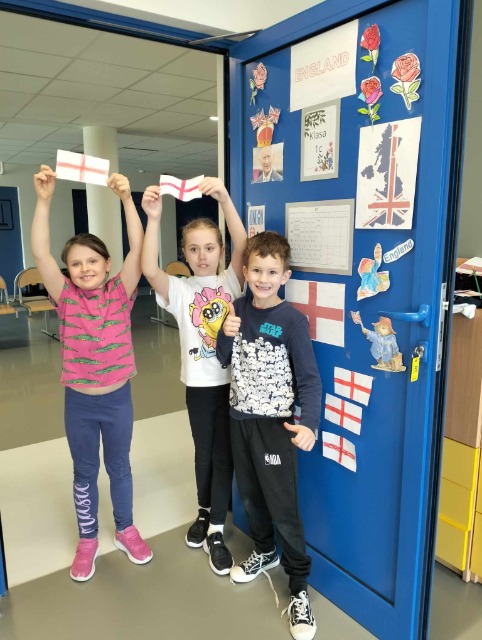 Trója dzieci, stojąca  przy otwartych drzwiach. Dwója dzieci trzyma w rękach zrobione z papieru angielskie flagi.