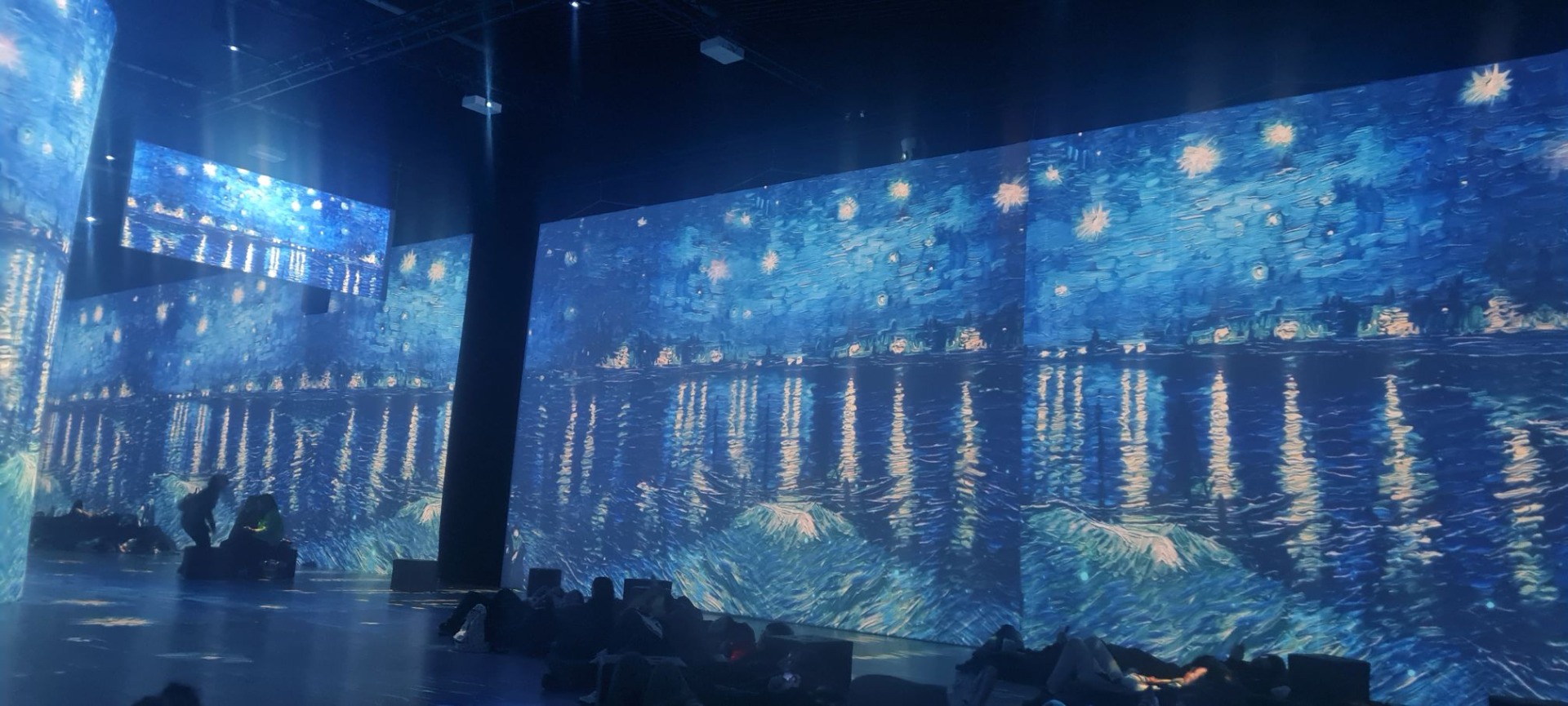 Obraz przedstawiający morze nocą wyświetlony na wielkoformatowej powierzchni. Widoczne sylwetki osób oglądających wystawę.