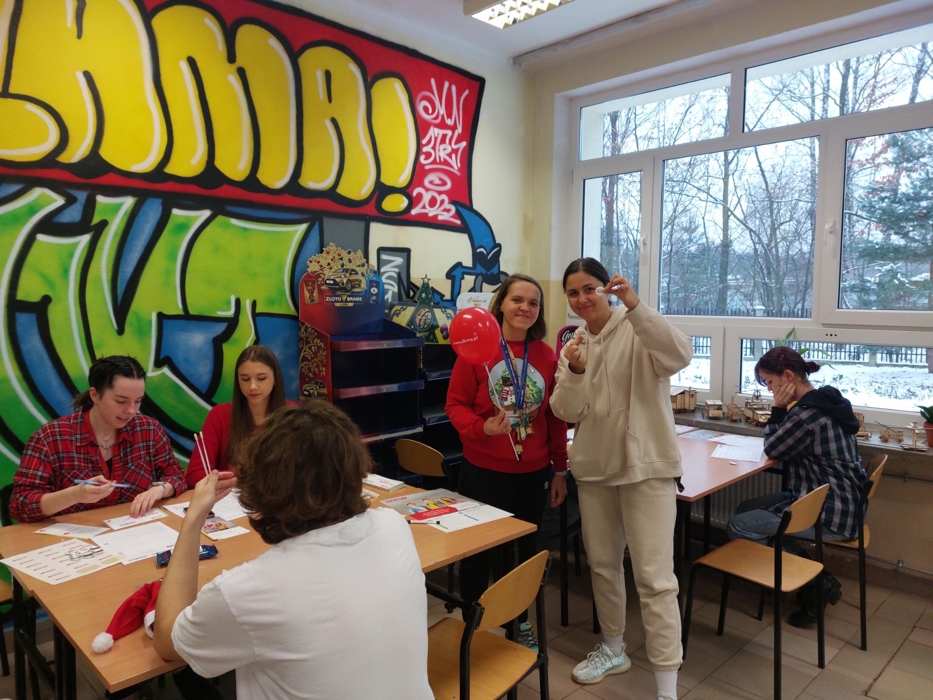 Wolontariuszki siedzące i obsługujące stoisko, obok dwie nauczycielki biorące udział w akcji stojące po prawej: Honorata Glazer i Karolina  Domagała. W tle graffiti na ścianie sali lekcyjnej. 
