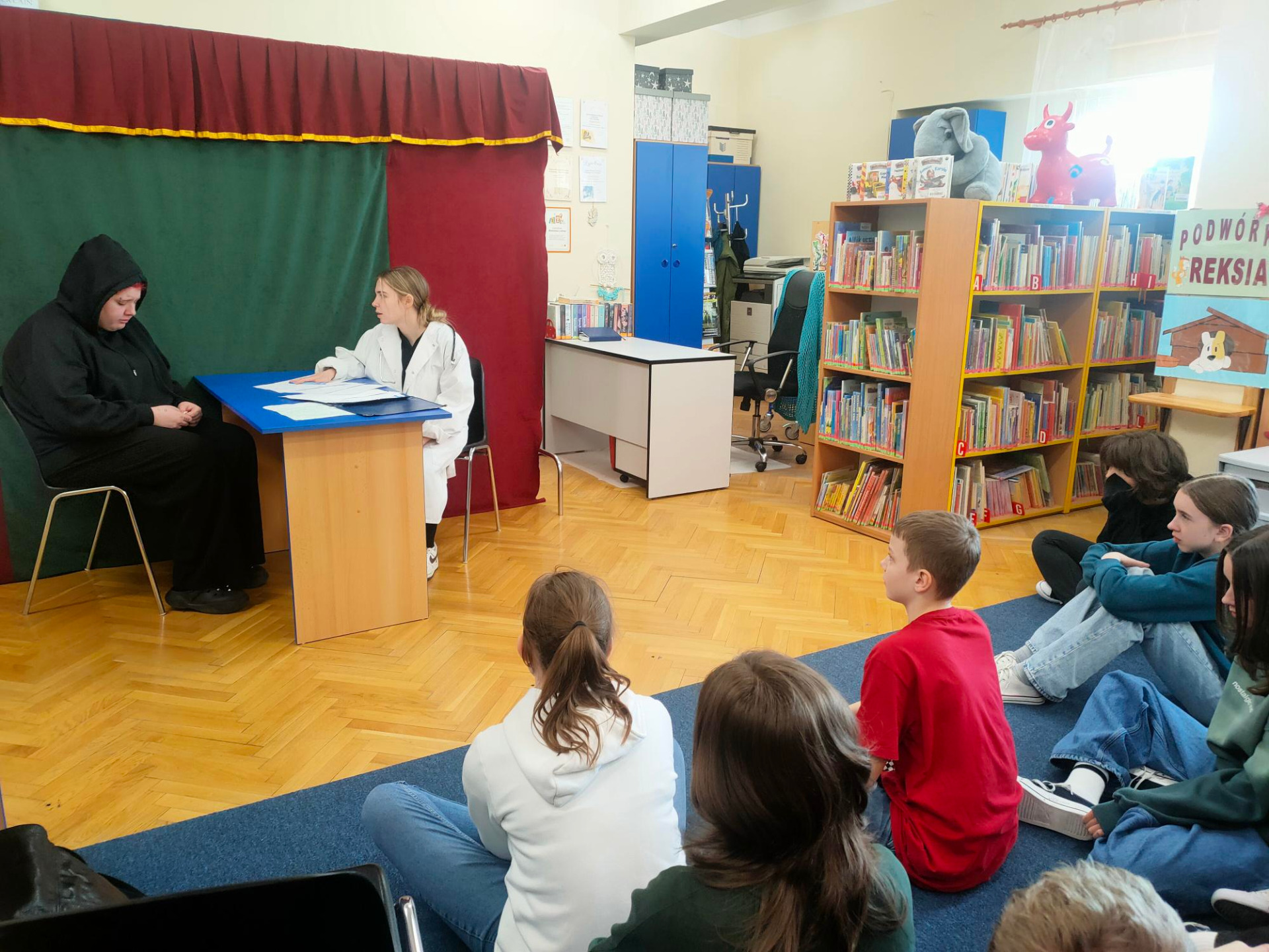 Uczniowie siedzą tyłem w bibliotece, przed nimi dwie postacie przy biurku