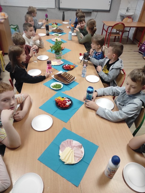 Jedenastu uczniów siedzi po dwóch stronach długiego stołu. Na stole znajdują się talerze oraz pokrojony chleb, warzywa, ser, wędlina i bazylią.