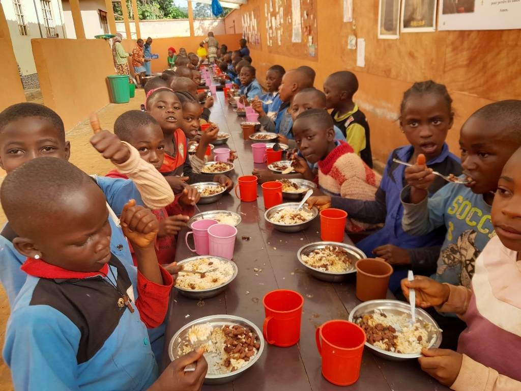 Zdjęcie przedstawia bardzo dużą grupę dzieci, które spożywają posiłek przy bardzo długim stole. Dzieci są ubrane w kolorowe ubrania. Na stole stoją metalowe talerze pełne jedzenia i kolorowe plastikowe kubki. Dzieci mają uśmiechnięte twarze.