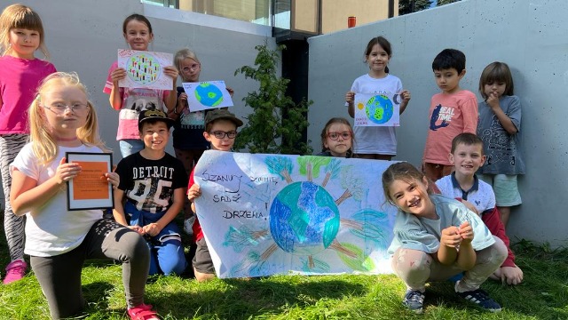 Kilkoro dzieci trzymające narysowane przez siebie plakaty z hasłami. Na jednym z nich można przeczytać "Szanuj zieleń, sadź zieleń".
Dzieci stoją wokół sadzonego przez nich drzewka.